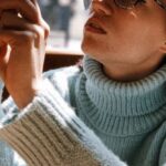 Knitwear Technology - Woman Wearing Light-green Turtleneck Sweater and Eyeglasses Using Phone While Sitting Near Brown Wooden Table Beside Glass Wall
