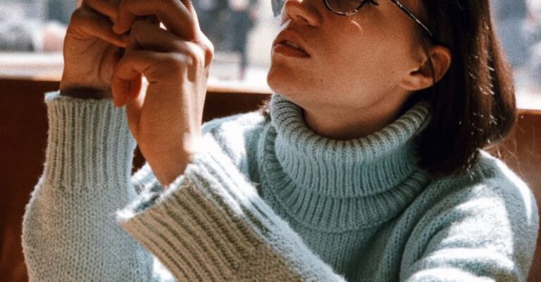 Knitwear Technology - Woman Wearing Light-green Turtleneck Sweater and Eyeglasses Using Phone While Sitting Near Brown Wooden Table Beside Glass Wall