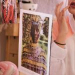Vintage Garments - Young stylish female in eyeglasses and turban sitting on bed in bedroom and adjusting makeup while looking in mirror