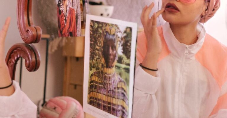 Vintage Garments - Young stylish female in eyeglasses and turban sitting on bed in bedroom and adjusting makeup while looking in mirror