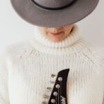 Plain Outfit - Unrecognizable female musician wearing white knitted sweater and stylish hat standing against white plain background with electric guitar fretboard in hands